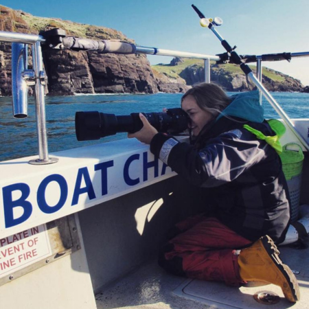 Dr Chiara Berteli is kneeling aboard a boat holding a camera up to her face to take a photograph.